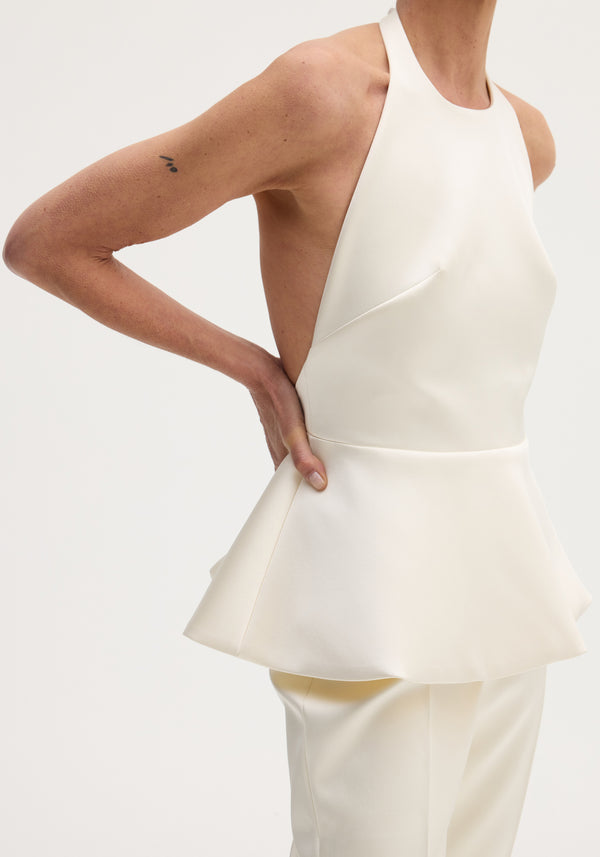Person wearing a Sculptural satin halter top | ivory, featuring a fitted bodice, flared peplum hem, and open sides, paired with matching trousers.