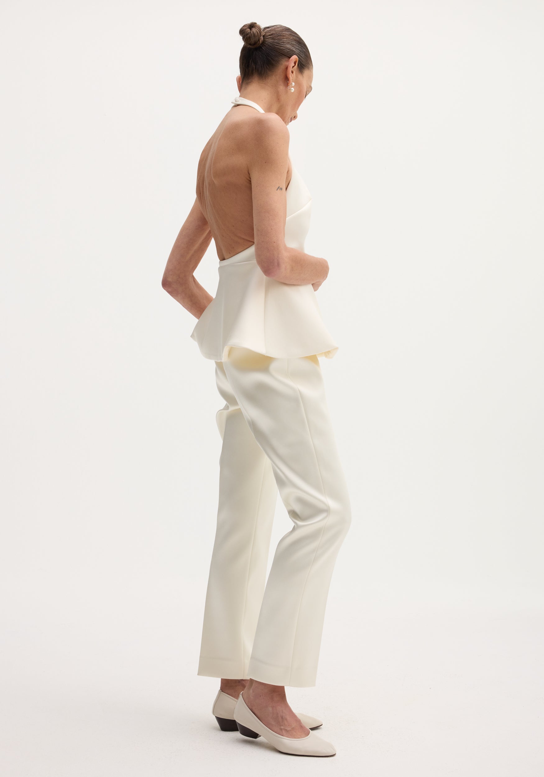 Slim woman in profile wearing a Sculptural satin halter top | ivory, with a low open back, peplum flare, and matching straight-leg trousers.