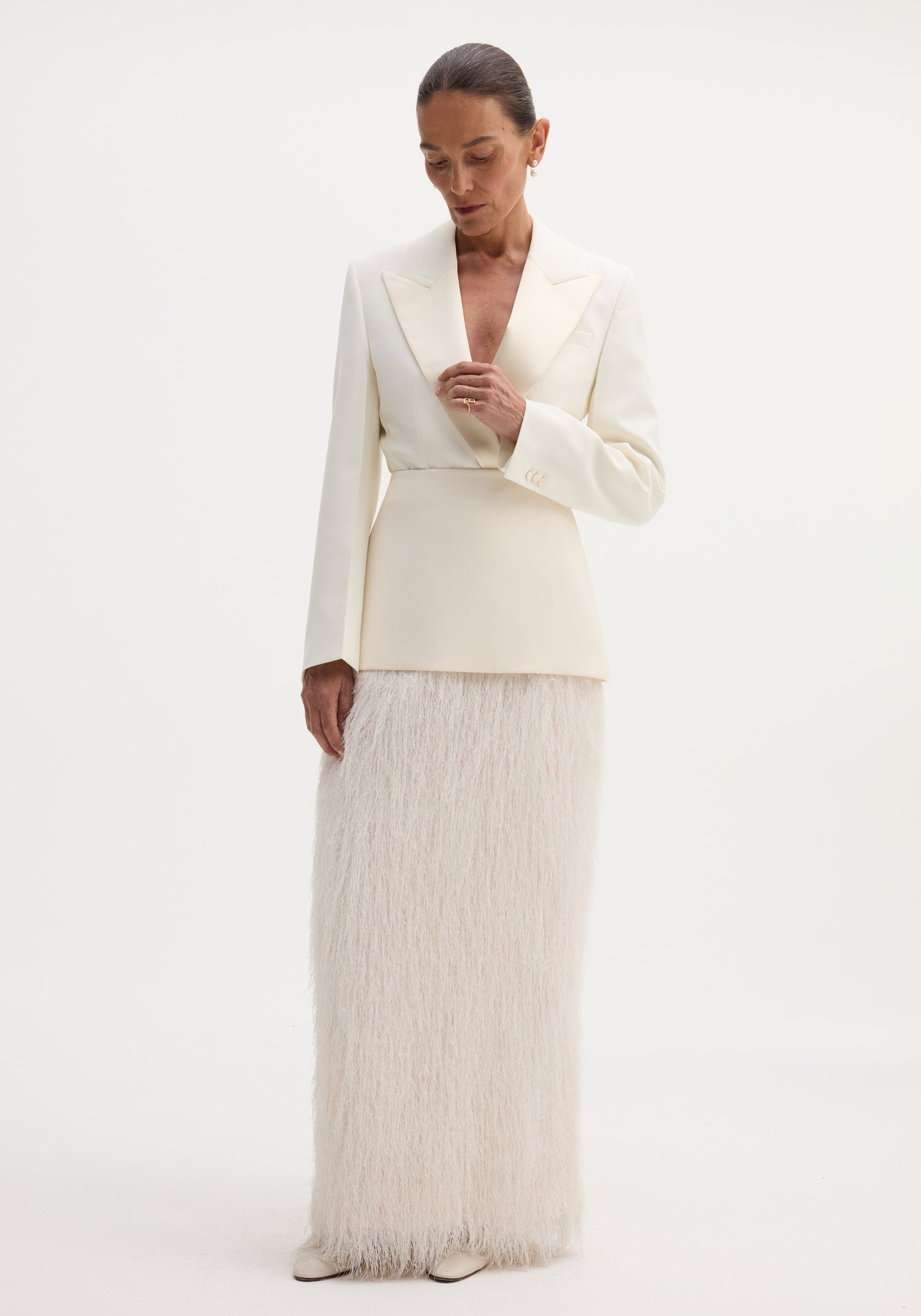 Woman wearing an ivory Fringe skirt with a structured wool upper and flowing fringe detail, touching her blazer lapel against a plain backdrop.