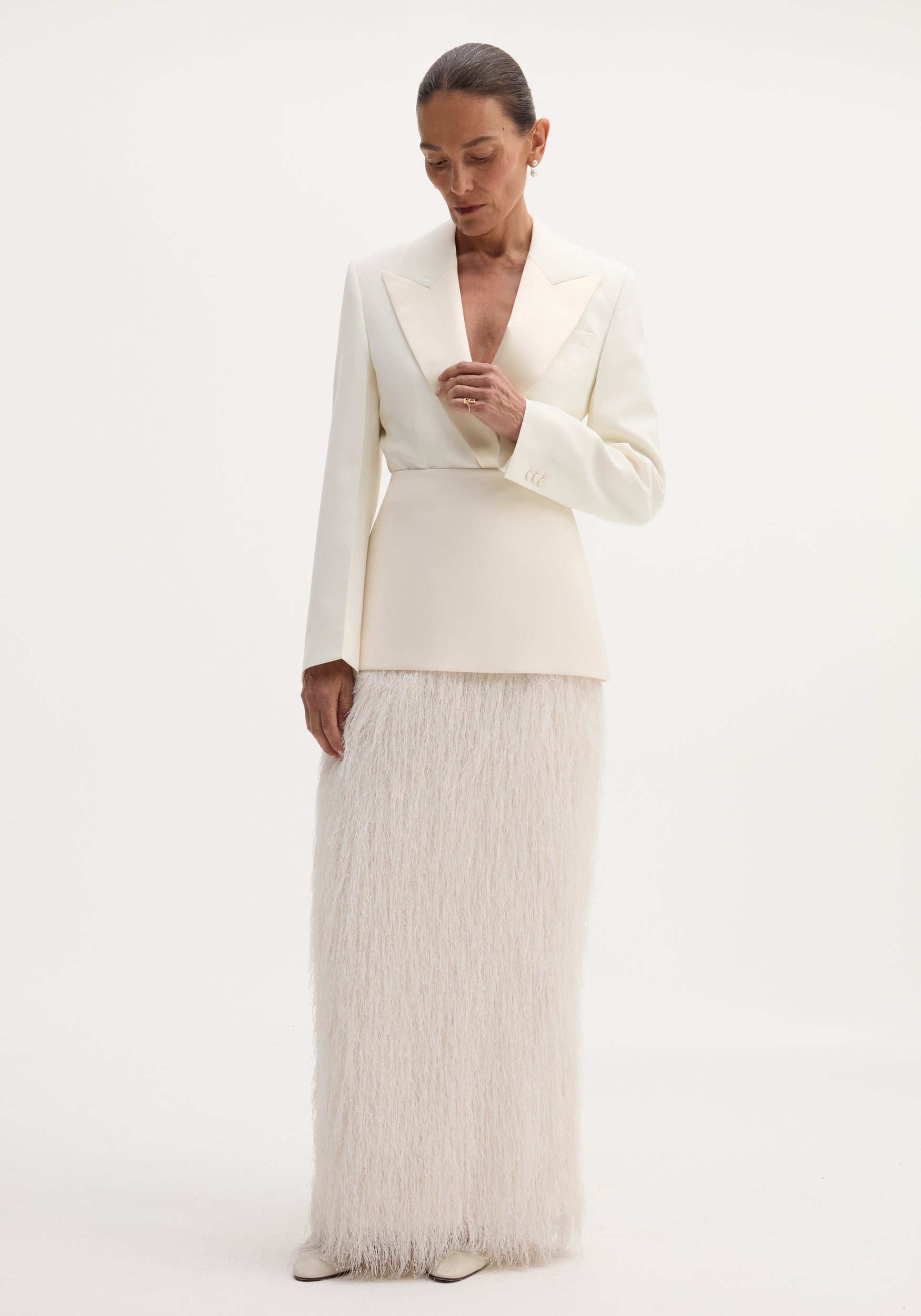 Woman wearing an ivory Fringe skirt with a structured wool upper and flowing fringe detail, touching her blazer lapel against a plain backdrop.