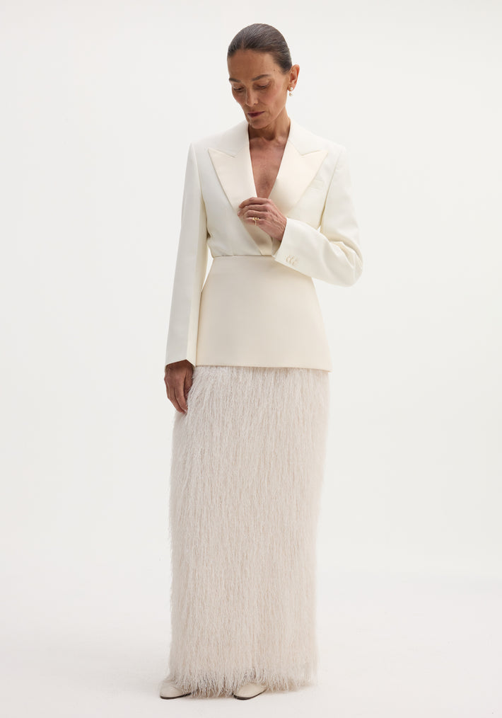 Woman wearing an ivory Fringe skirt with a structured wool upper and flowing fringe detail, touching her blazer lapel against a plain backdrop.