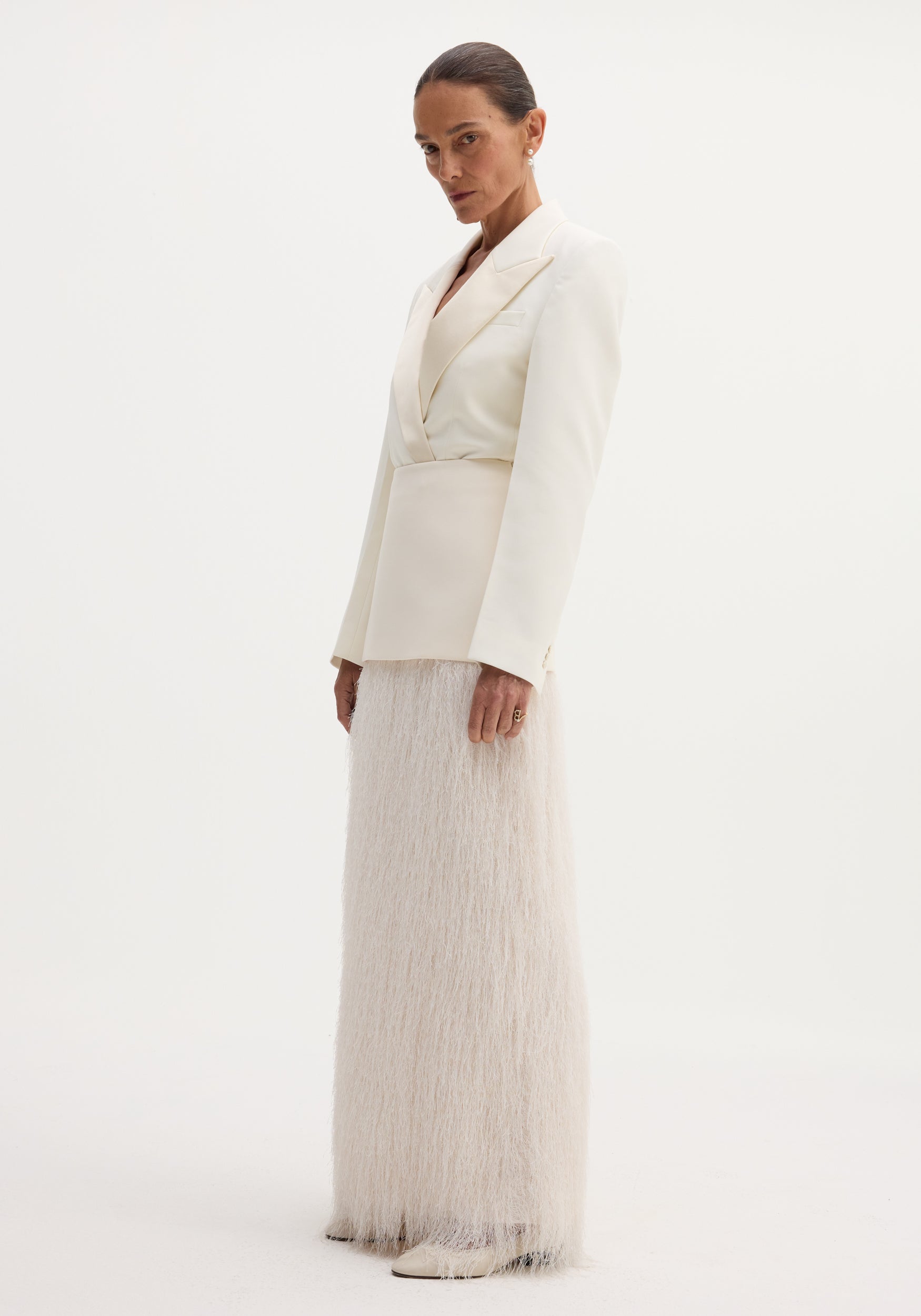 Model wearing Fringe skirt in ivory with floor-length fringe detail, tailored blazer, drop pearl earrings, and point-toed shoes, showcasing minimalist elegance.