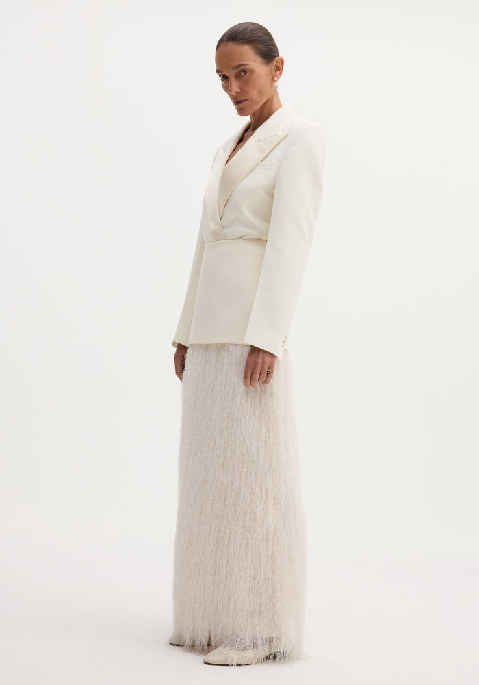 Model wearing Fringe skirt in ivory with floor-length fringe detail, tailored blazer, drop pearl earrings, and point-toed shoes, showcasing minimalist elegance.