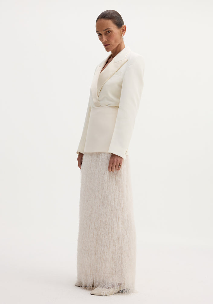 Woman wearing an ivory Fringe skirt with a structured wool upper and flowing fringe detail, touching her blazer lapel against a plain backdrop.