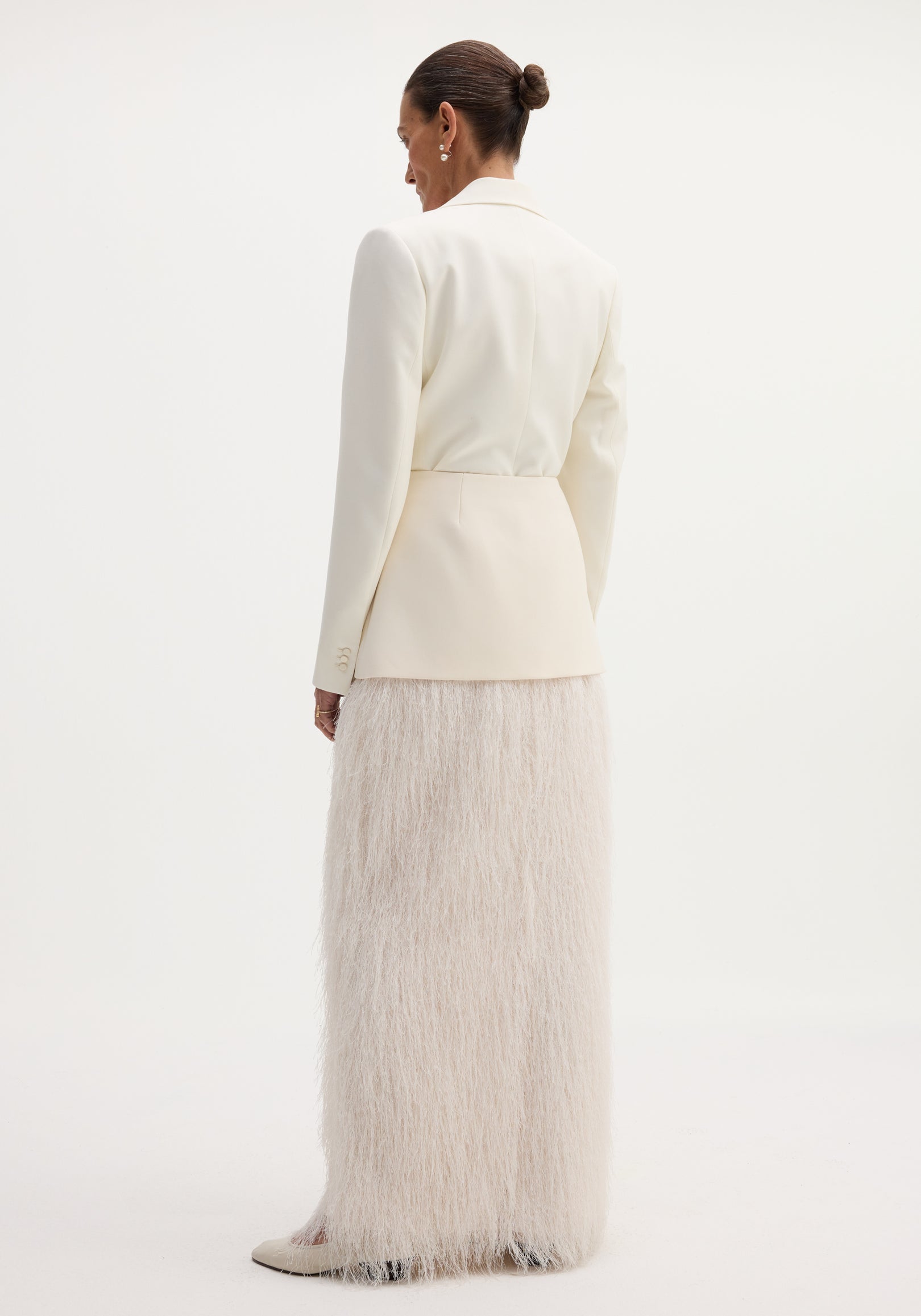 Model in off-white blazer showcasing the Fringe skirt | ivory, featuring a fitted wool upper and full-length fringe, with pointed-toe heels visible.