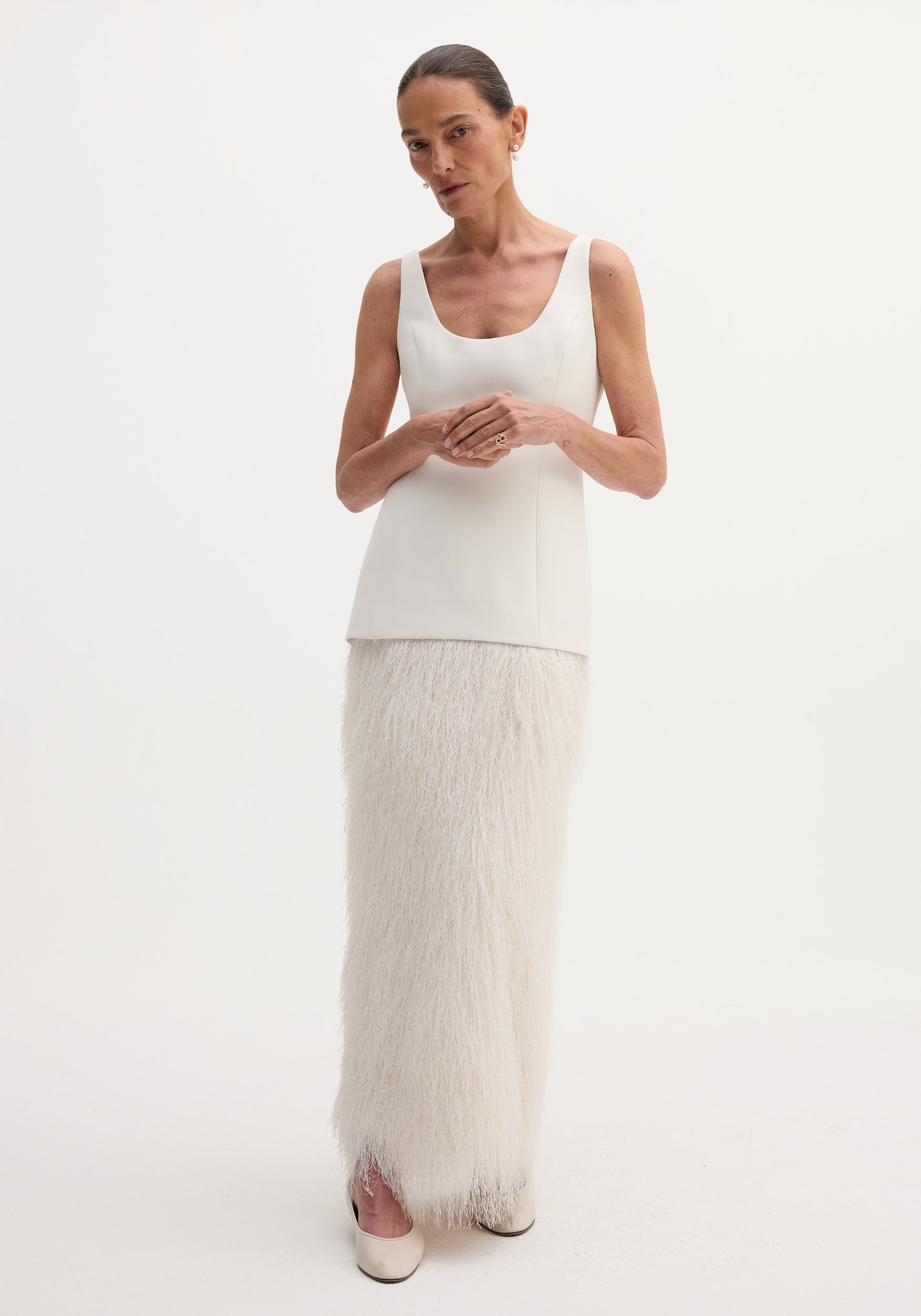 Woman models Fringe dress | ivory with a structured fused wool bodice and floor-length feathery fringe skirt, accessorized with pearl earrings and rings.