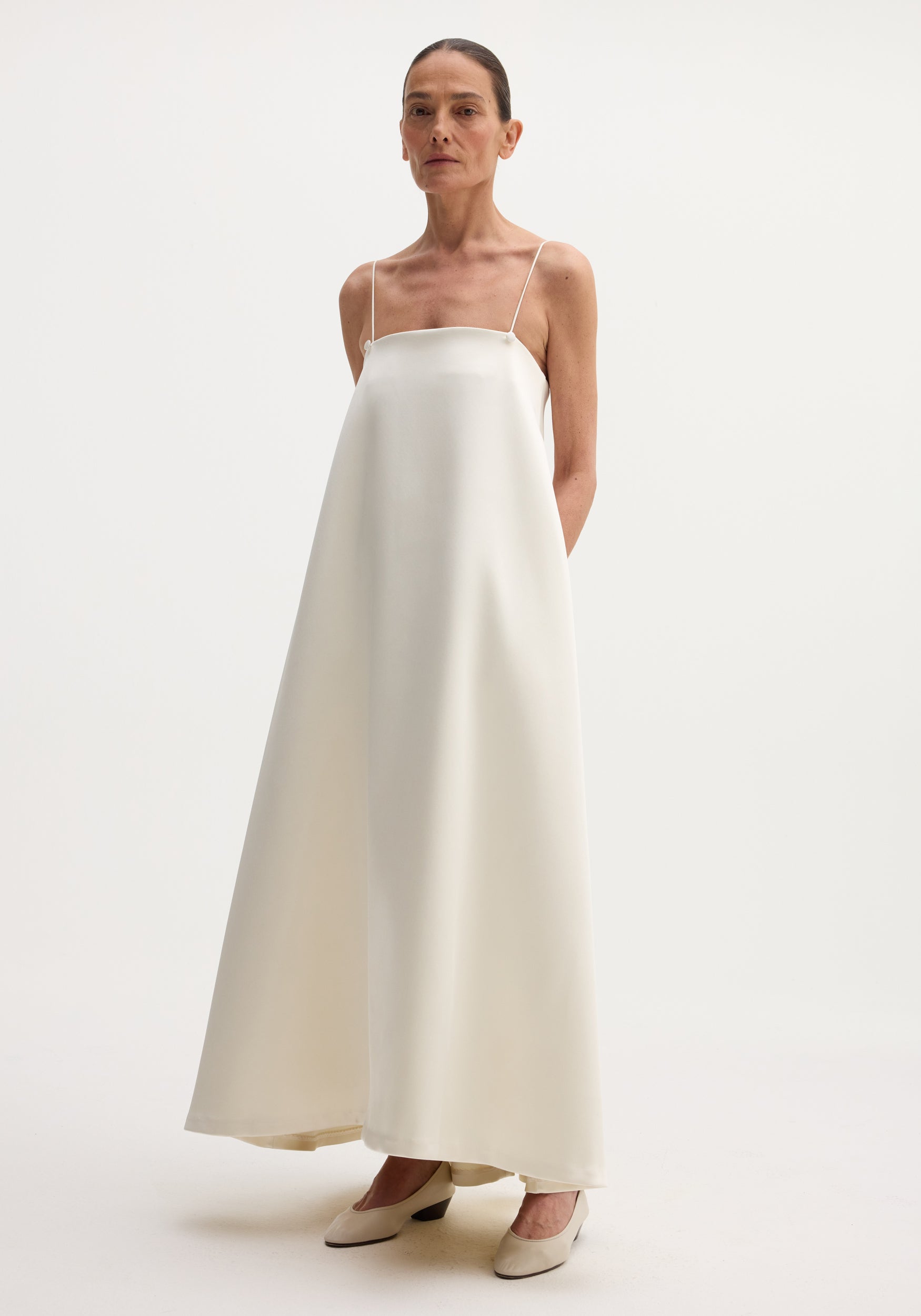 Voluminous satin dress | ivory, featuring a floor-length, loose A-line silhouette with adjustable thin straps, button detailing, and a straight neckline. Model pairs it with neutral pumps.