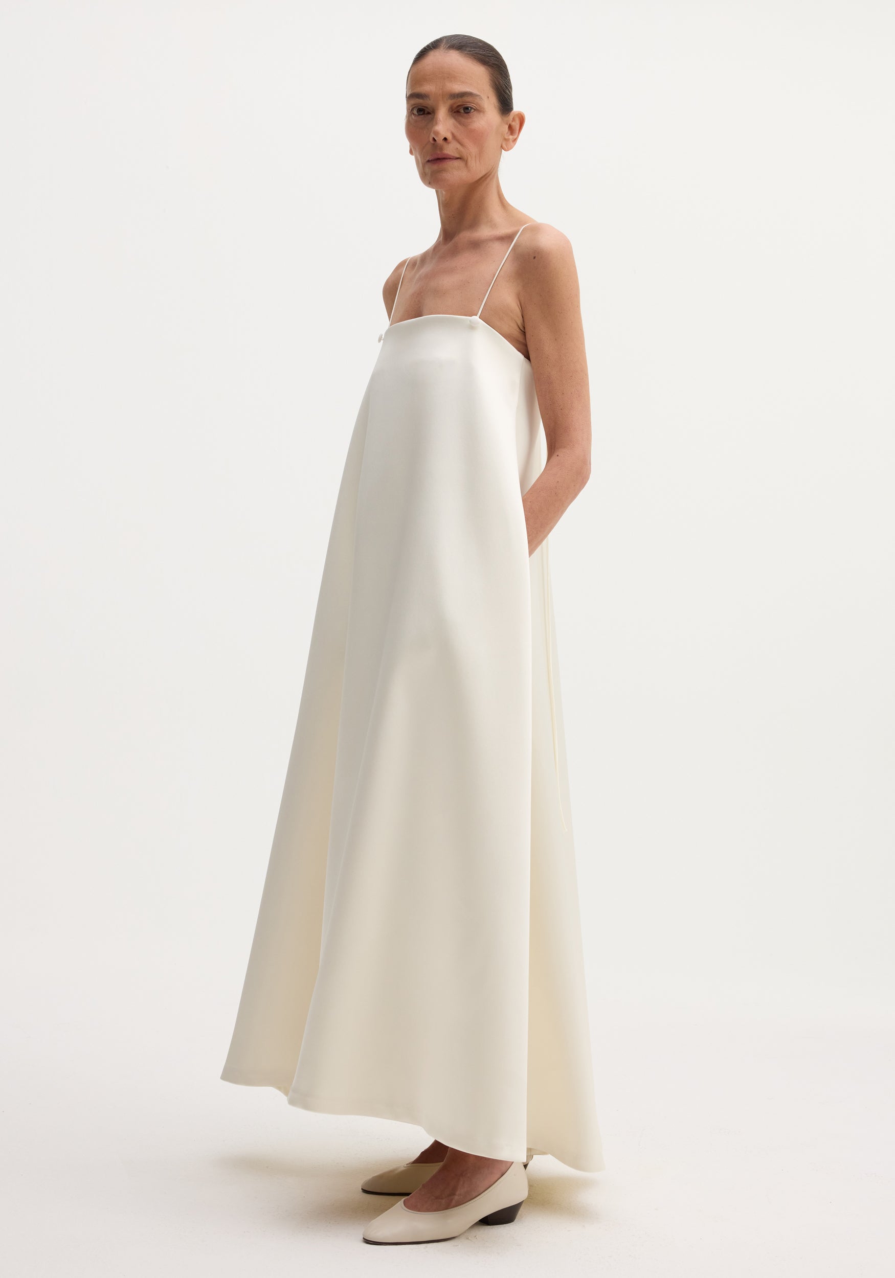 Model wearing Voluminous satin dress | ivory, featuring a full-length, flowy silhouette, adjustable spaghetti straps, and button details, with a modern, minimalist appeal.