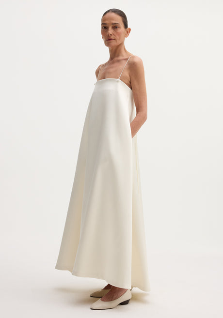 Voluminous satin dress | ivory, featuring a floor-length, loose A-line silhouette with adjustable thin straps, button detailing, and a straight neckline. Model pairs it with neutral pumps.