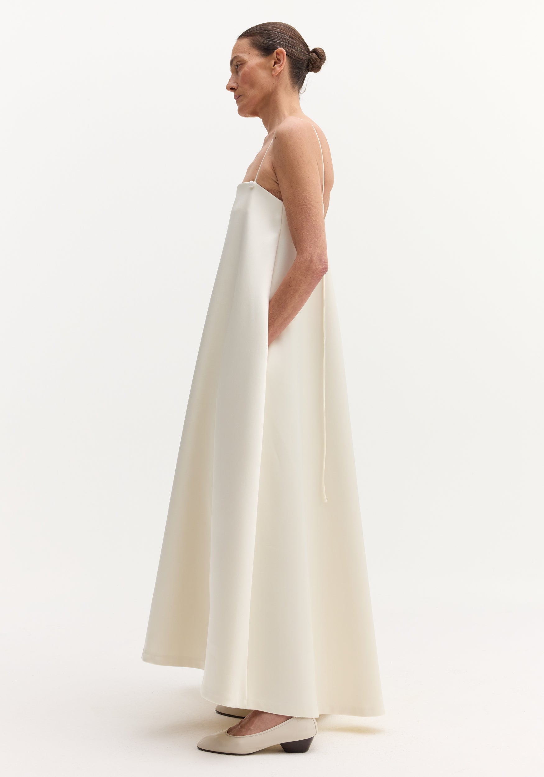 Side profile of a woman in a Voluminous satin dress | ivory with adjustable spaghetti straps, open back, and hidden pockets, styled with a neat bun.