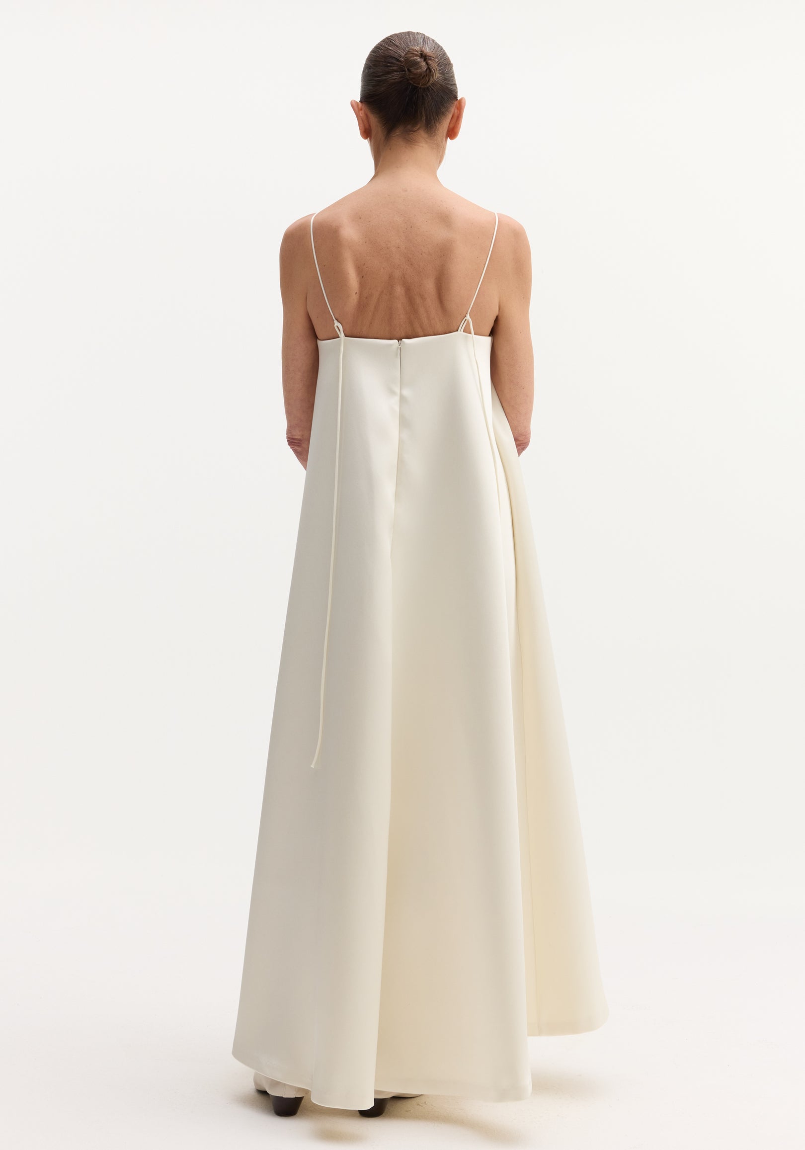 Woman in Voluminous satin dress | ivory, seen from behind, wearing a floor-length gown with adjustable spaghetti straps tying at the back, and button details.