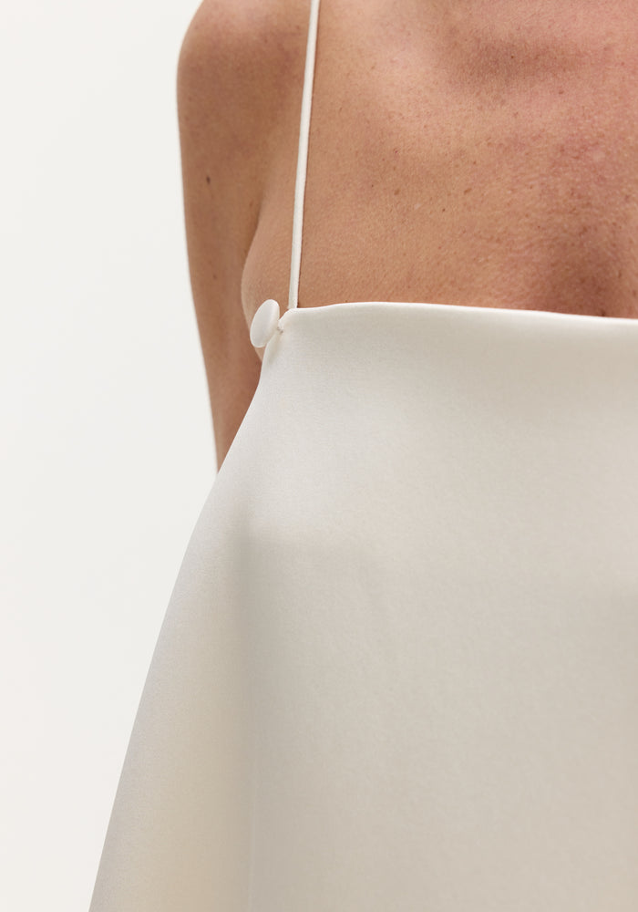 Close-up of a model wearing an ivory Voluminous satin dress, showcasing its silky texture, thin adjustable straps with button detail, and smooth drape across the chest.