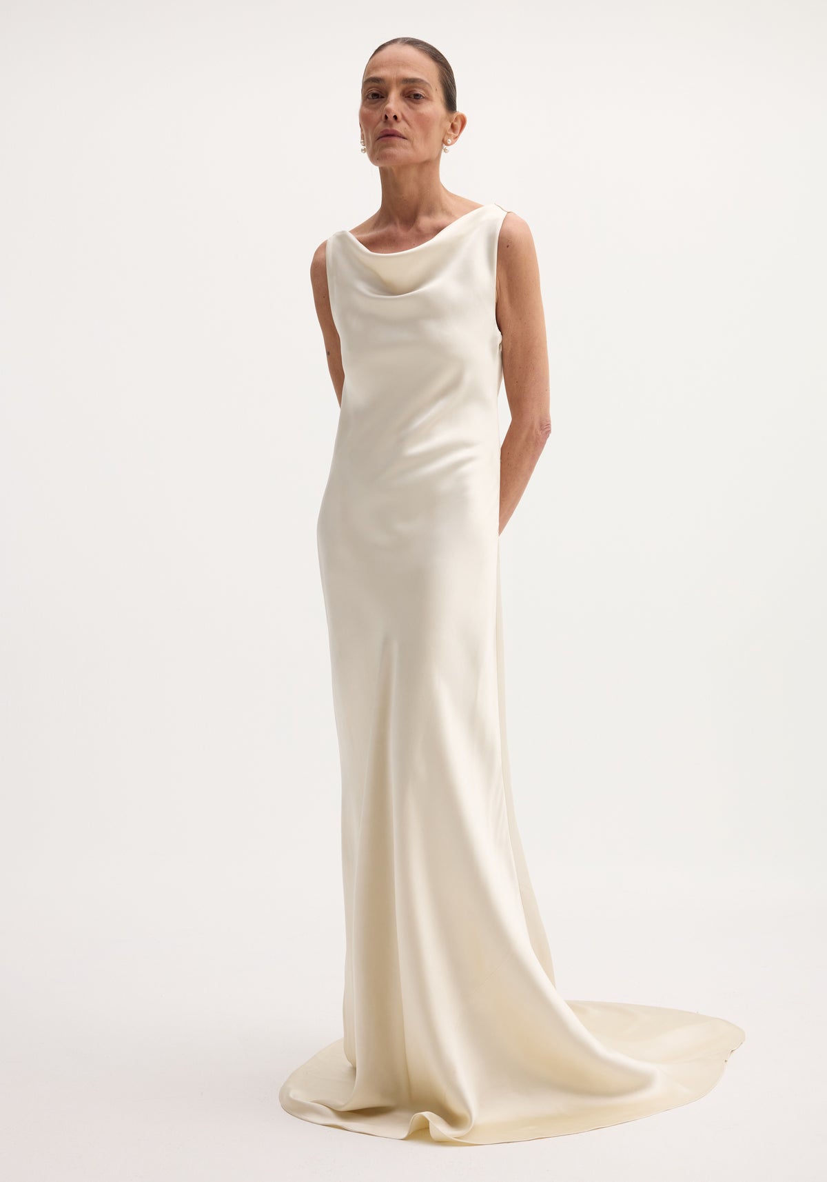 Tall model in an ivory Draped satin dress with train, featuring a cowl neckline, sleeveless design, and elegant drape, accessorized with pearl earrings.