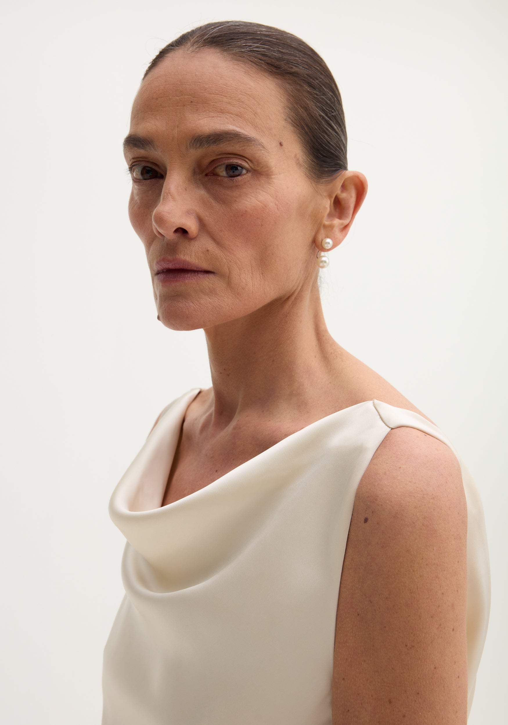 Woman in a sleeveless, draped satin dress with train, featuring a scoop neck and back, accessorized with drop pearl earrings.