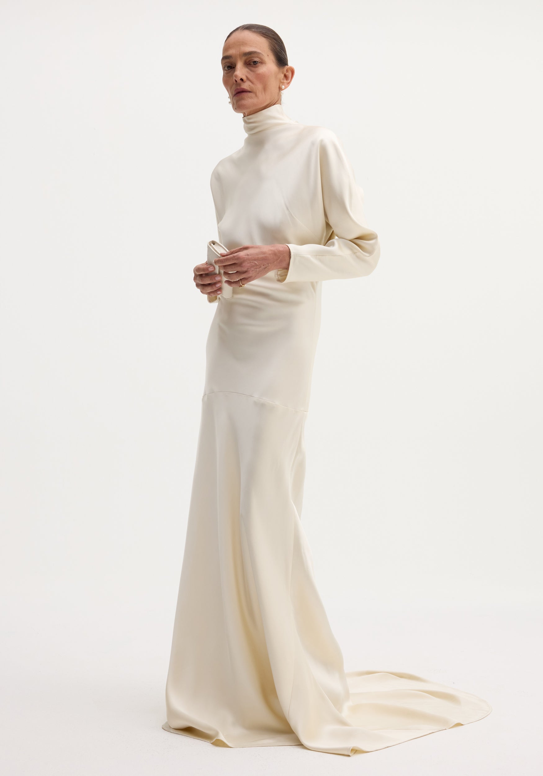 Model wearing Satin button down dress with train in ivory, featuring long sleeves, high neck, bias draping, and button closures on the back.