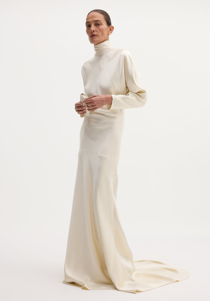 Model wearing Satin button down dress with train in ivory, featuring long sleeves, high neck, bias draping, and button closures on the back.