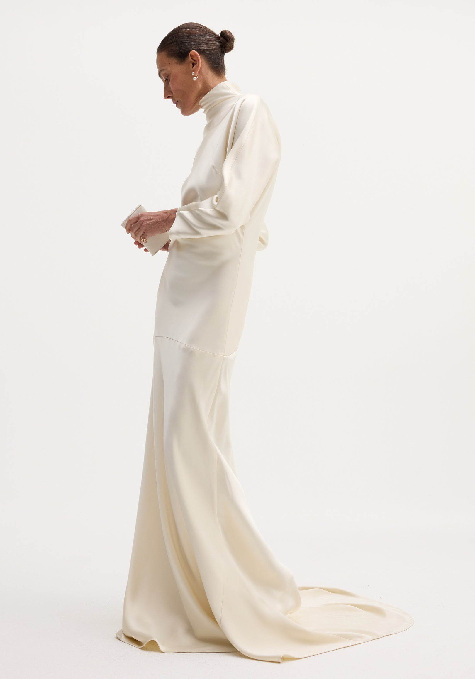Woman in profile wearing an ivory satin button-down dress with long sleeves, high neck, bias draping, and elegant train, holding a matching clutch.