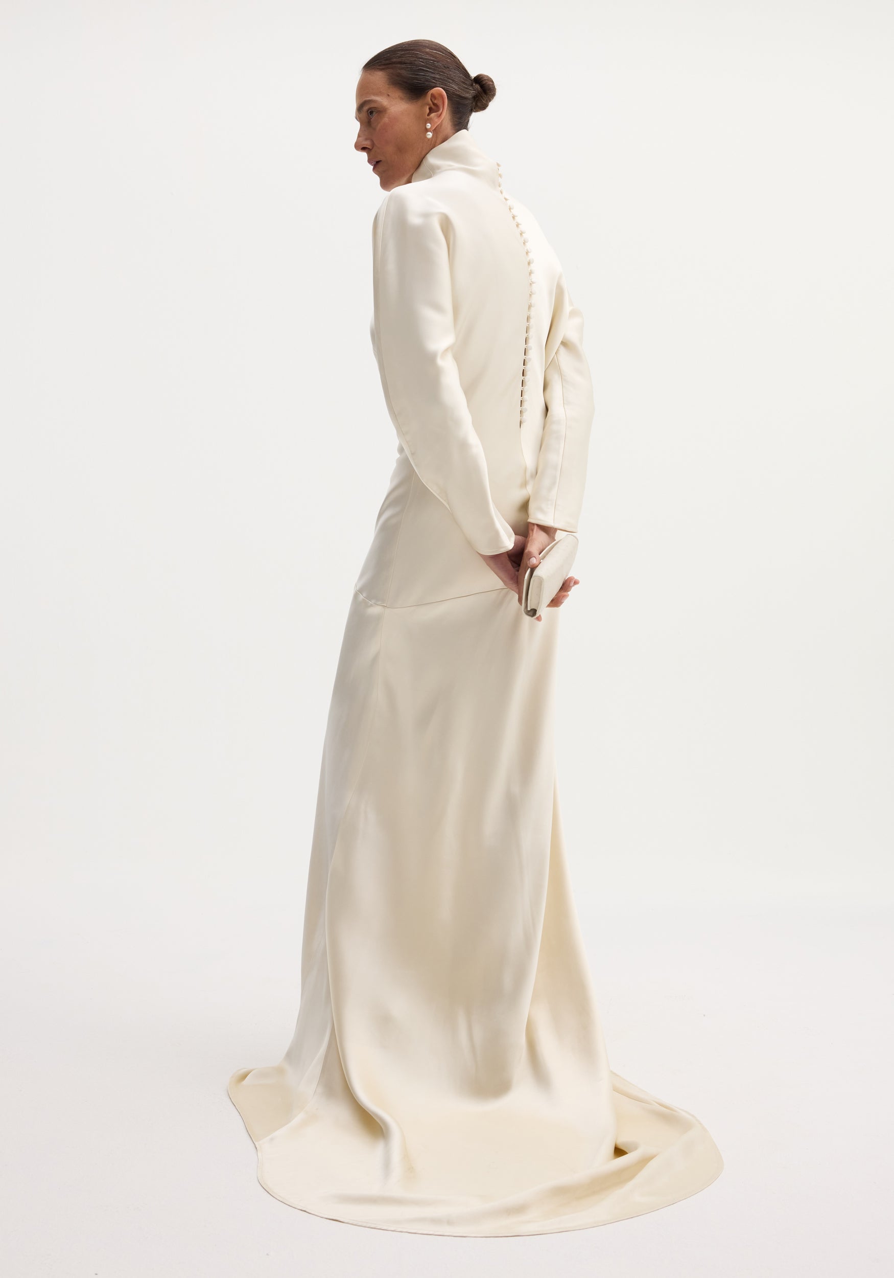 Person in profile wearing a Satin button down dress with train | ivory, featuring long sleeves, high neck, buttoned back, and holding a matching clutch.