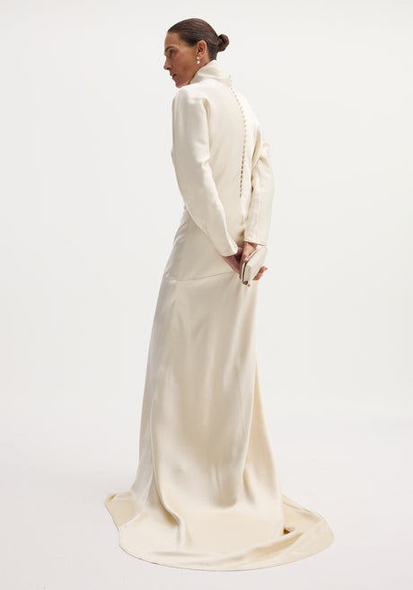 Model wearing Satin button down dress with train in ivory, featuring long sleeves, high neck, bias draping, and button closures on the back.