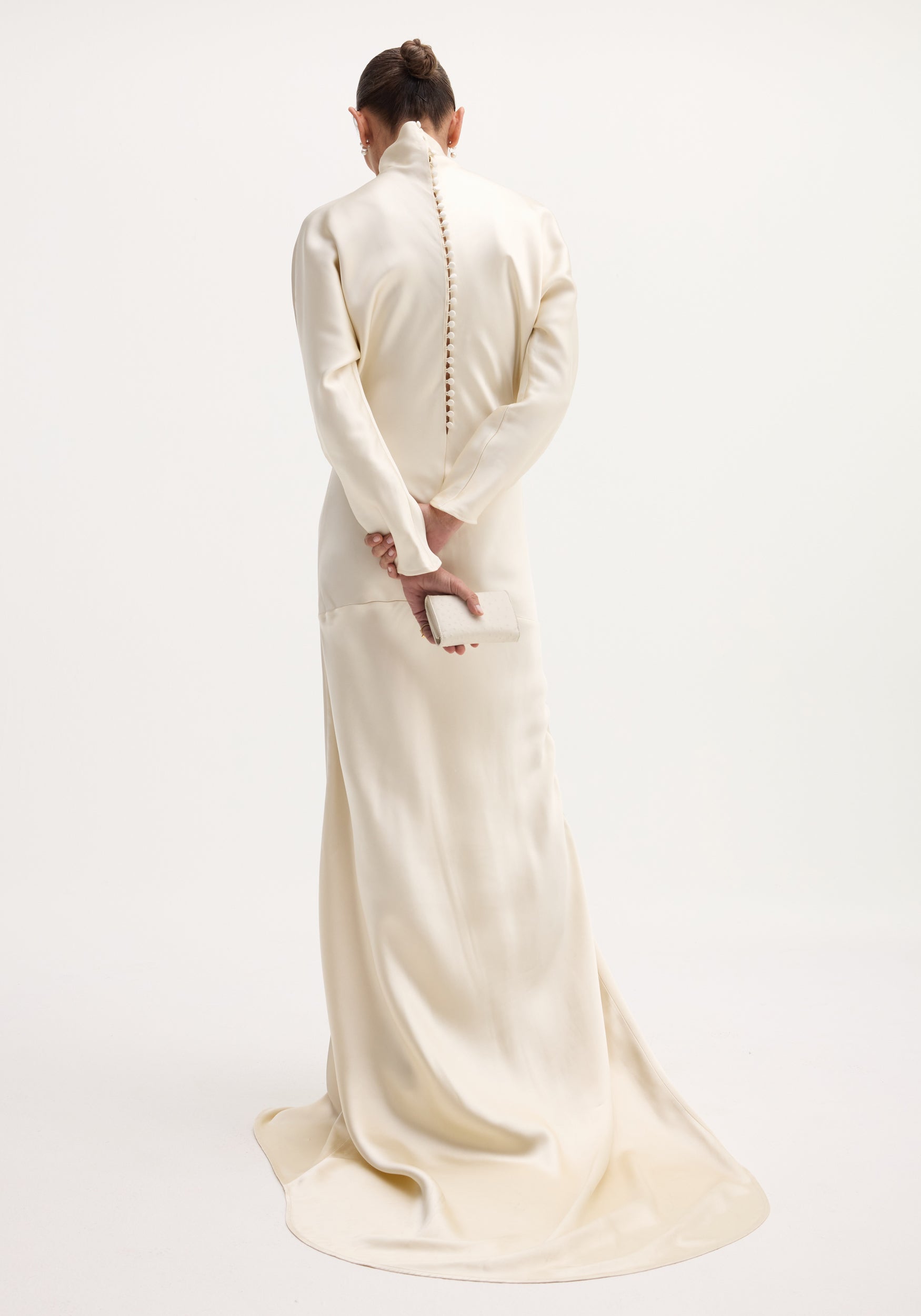 Person in ivory Satin button down dress with train, high collar, long sleeves, covered buttons down back, holding a matching clutch, elegant train pooling on floor.