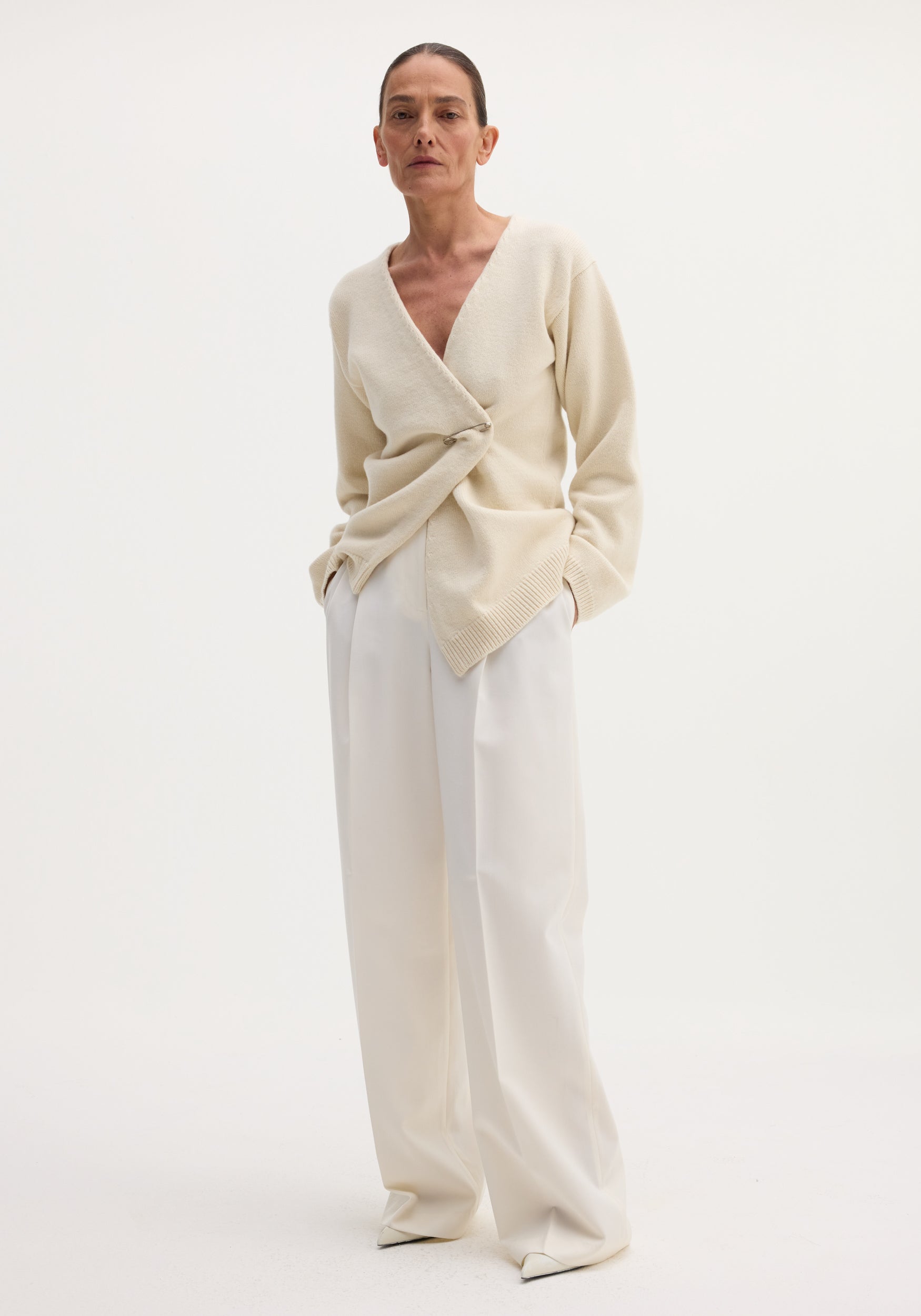 Off-shoulder wrap cardigan in cream, worn by a slender model, features a relaxed fit and metal safety-pin closure, paired with wide-leg trousers.