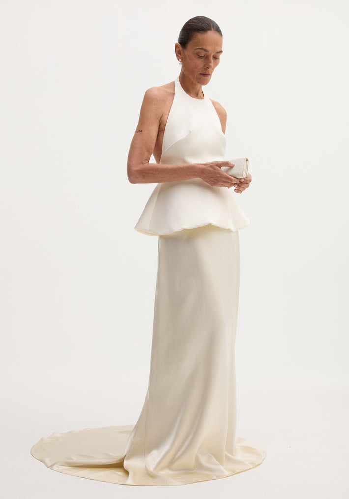 Woman wearing an ivory Sculptural satin halter top with separate silk skirt, featuring a peplum waist and flowing train, holding a matching clutch.