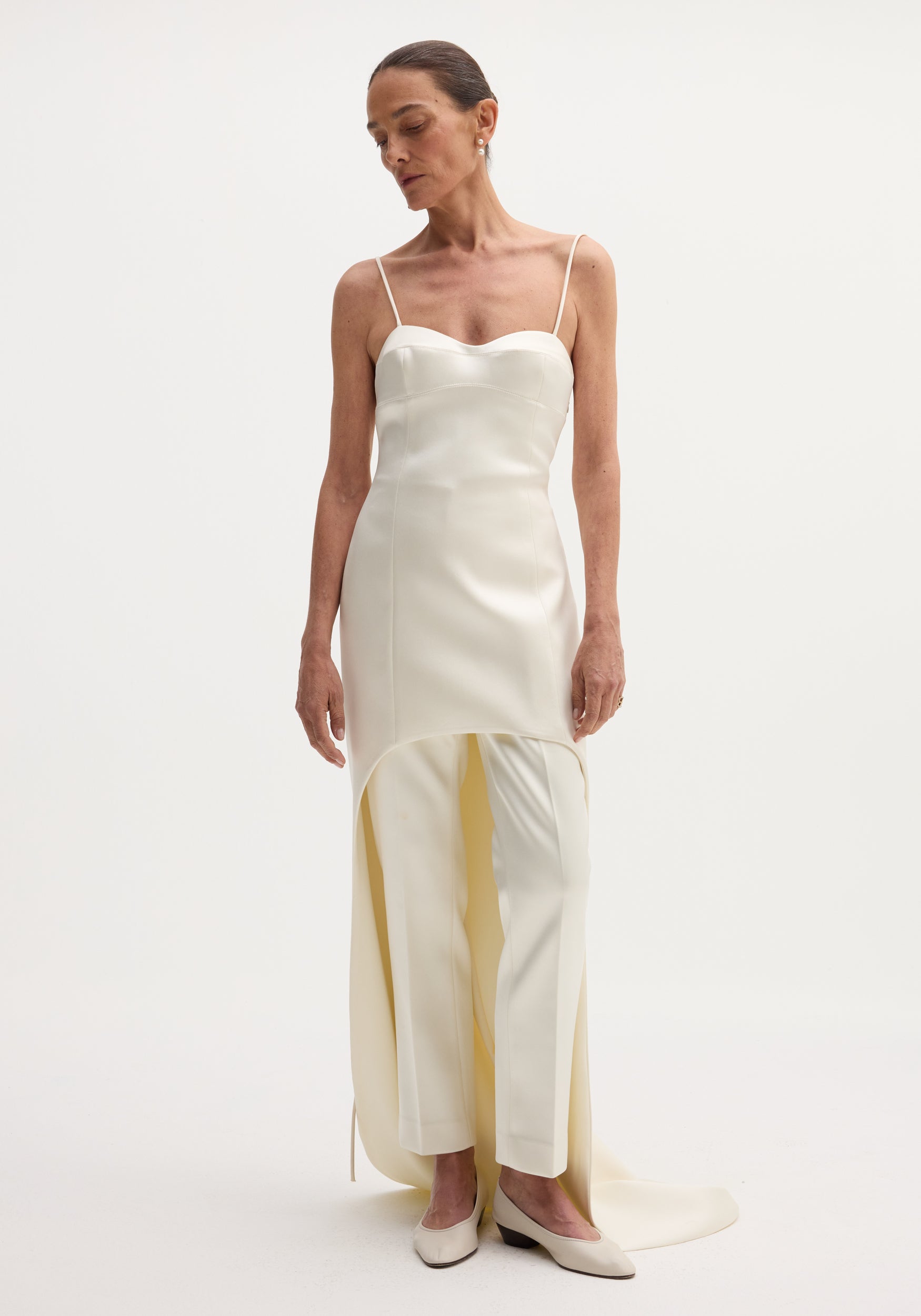 Model wearing Satin bustier dress with train, featuring a sleeveless bodice, asymmetrical overskirt, paired with straight-leg trousers, and accessorized with simple drop earrings.