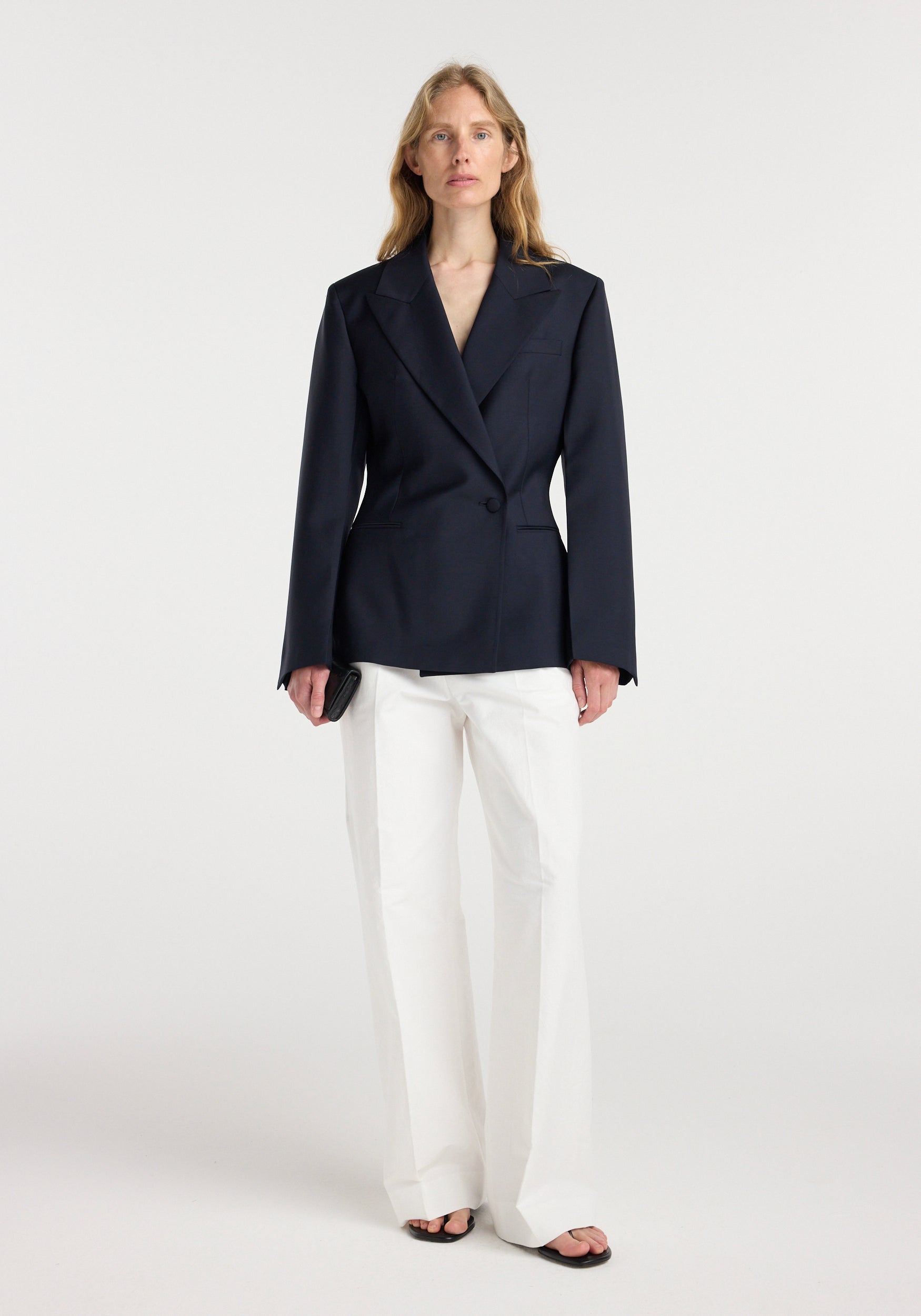 Woman wearing an hourglass shaped blazer | navy with a single button closure, paired with white wide-leg trousers and black open-toe sandals, standing against a plain background.