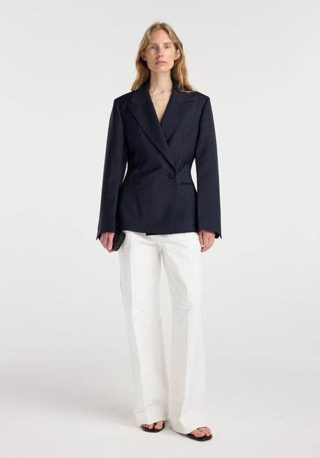 Woman wearing an hourglass shaped blazer | navy with a single button closure, paired with white wide-leg trousers and black open-toe sandals, standing against a plain background.