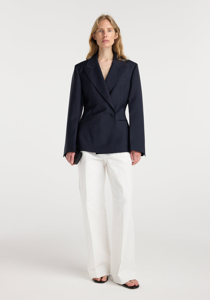 Woman wearing an hourglass shaped blazer | navy with a single button closure, paired with white wide-leg trousers and black open-toe sandals, standing against a plain background.