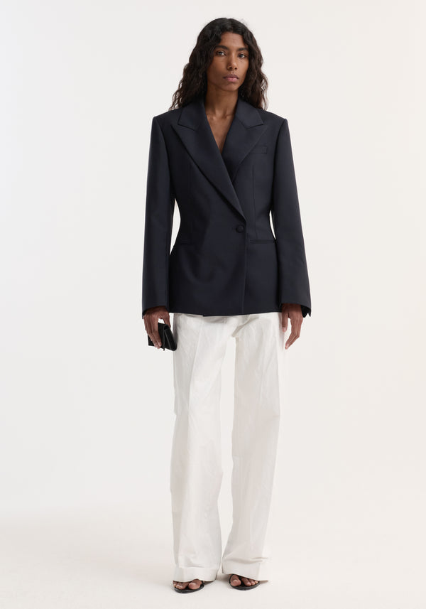 Woman wearing Hourglass shaped blazer with sharp notch lapel, cinched waist, single button closure, paired with white trousers and holding a black clutch.