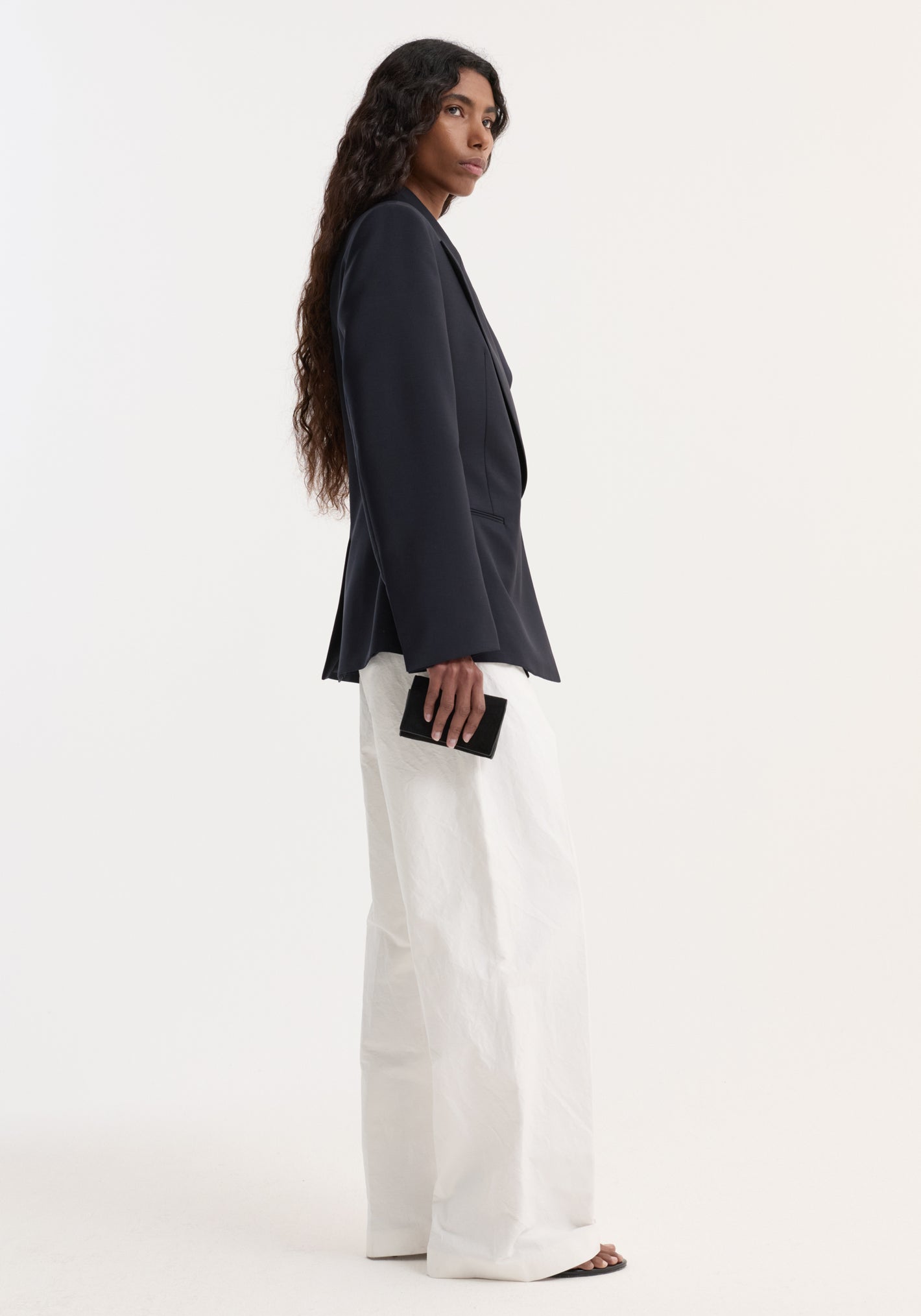 Person in a tailored Hourglass shaped blazer with sharp lapels, cinched waist, over white trousers, holding a black clutch, showcasing elegant, architectural tailoring.