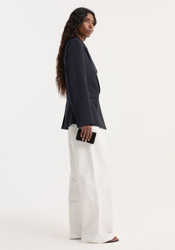 Person in a tailored Hourglass shaped blazer with sharp lapels, cinched waist, over white trousers, holding a black clutch, showcasing elegant, architectural tailoring.
