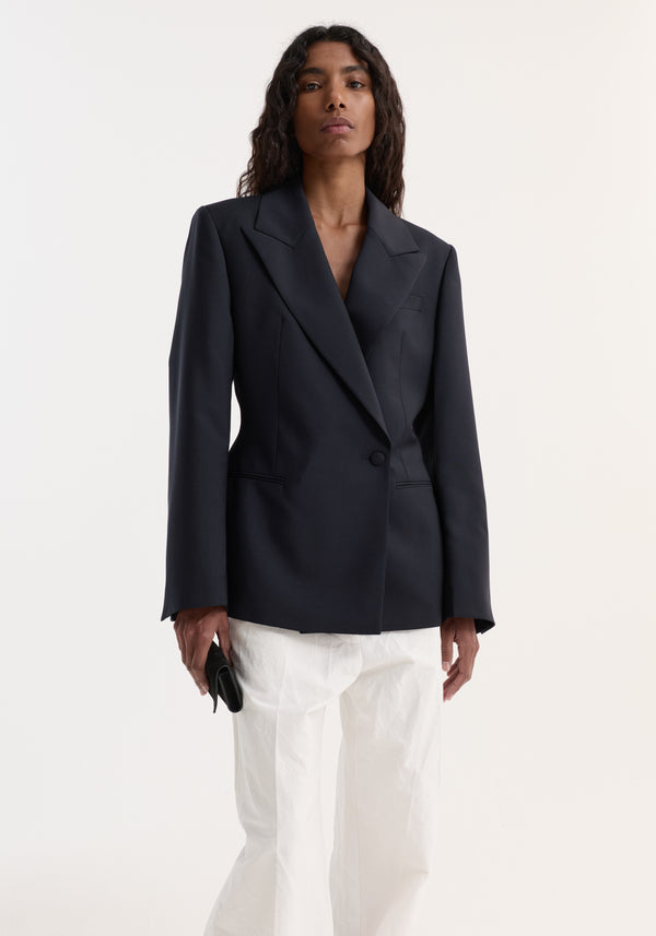 Model in Hourglass shaped blazer with structured shoulders, notch lapel, and cinched waist, paired with loose white trousers, holding a black accessory.