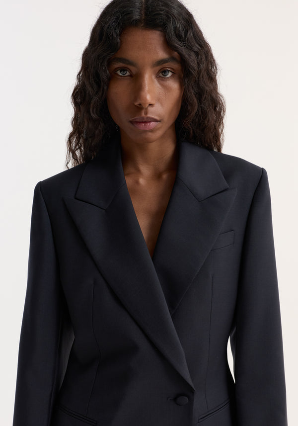 Person wearing Hourglass shaped blazer with sharp notch lapel, cinched waist, and single button closure. Tailored fit highlights architectural silhouette and defined shoulders.