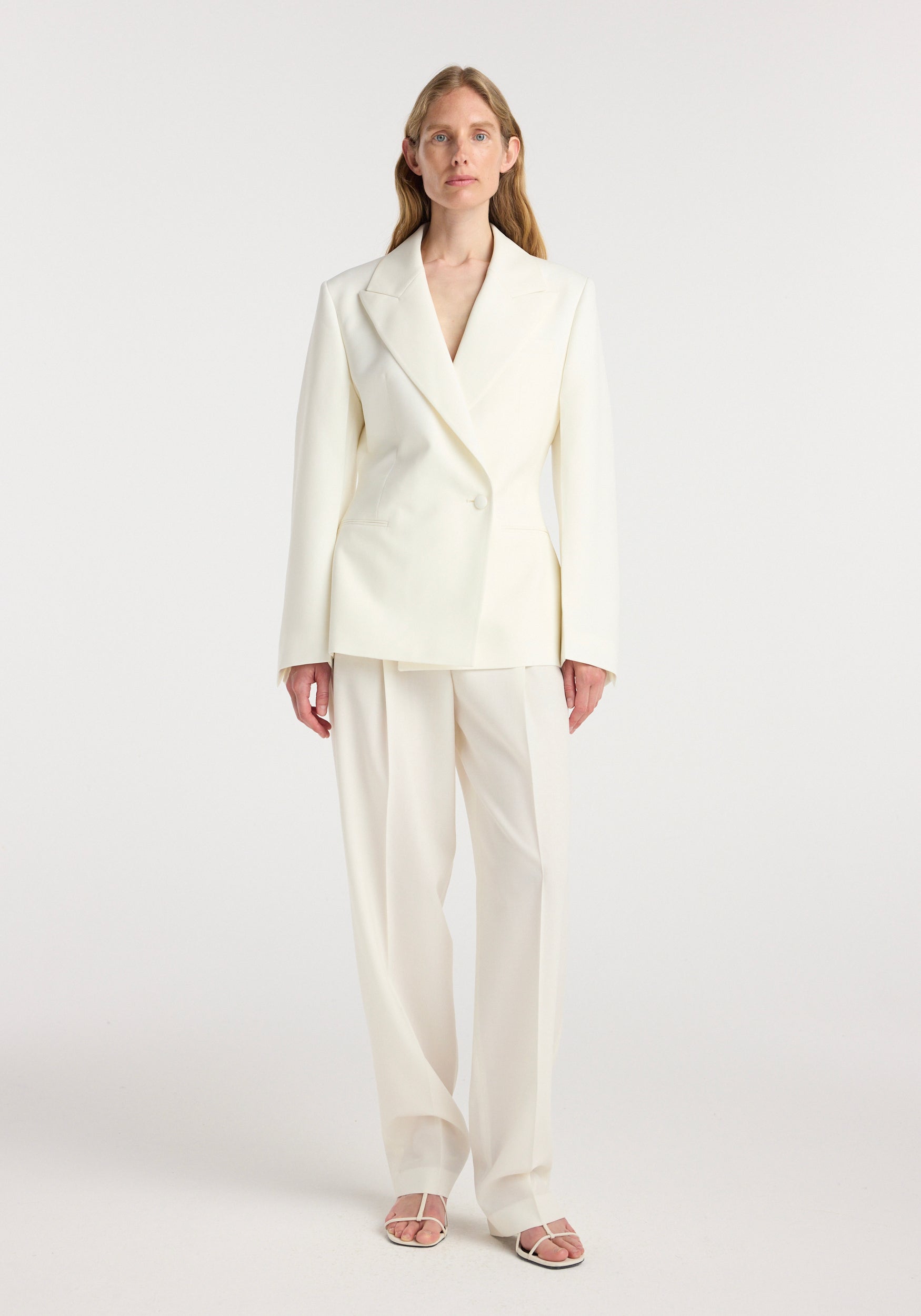 Woman wearing an off white hourglass shaped blazer with tailored lapels and a single button closure, paired with matching wide-leg trousers and open-toe sandals.