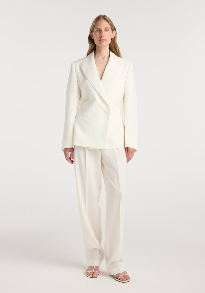 Woman wearing an off white hourglass shaped blazer with tailored lapels and a single button closure, paired with matching wide-leg trousers and open-toe sandals.