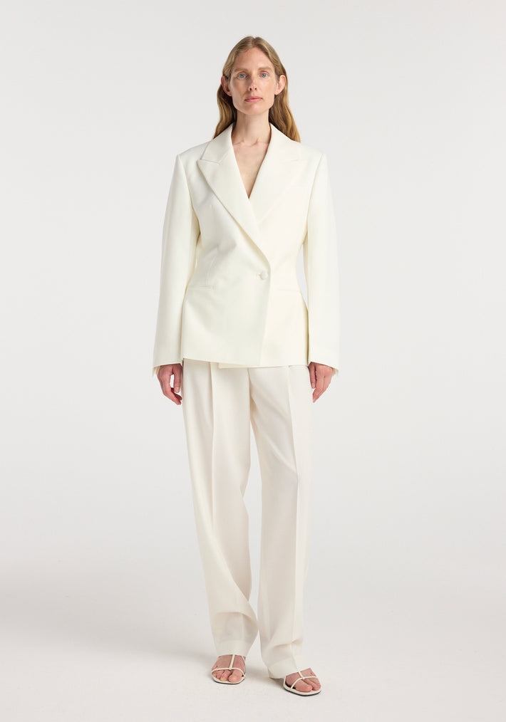 Woman wearing an off white hourglass shaped blazer with tailored lapels and a single button closure, paired with matching wide-leg trousers and open-toe sandals.