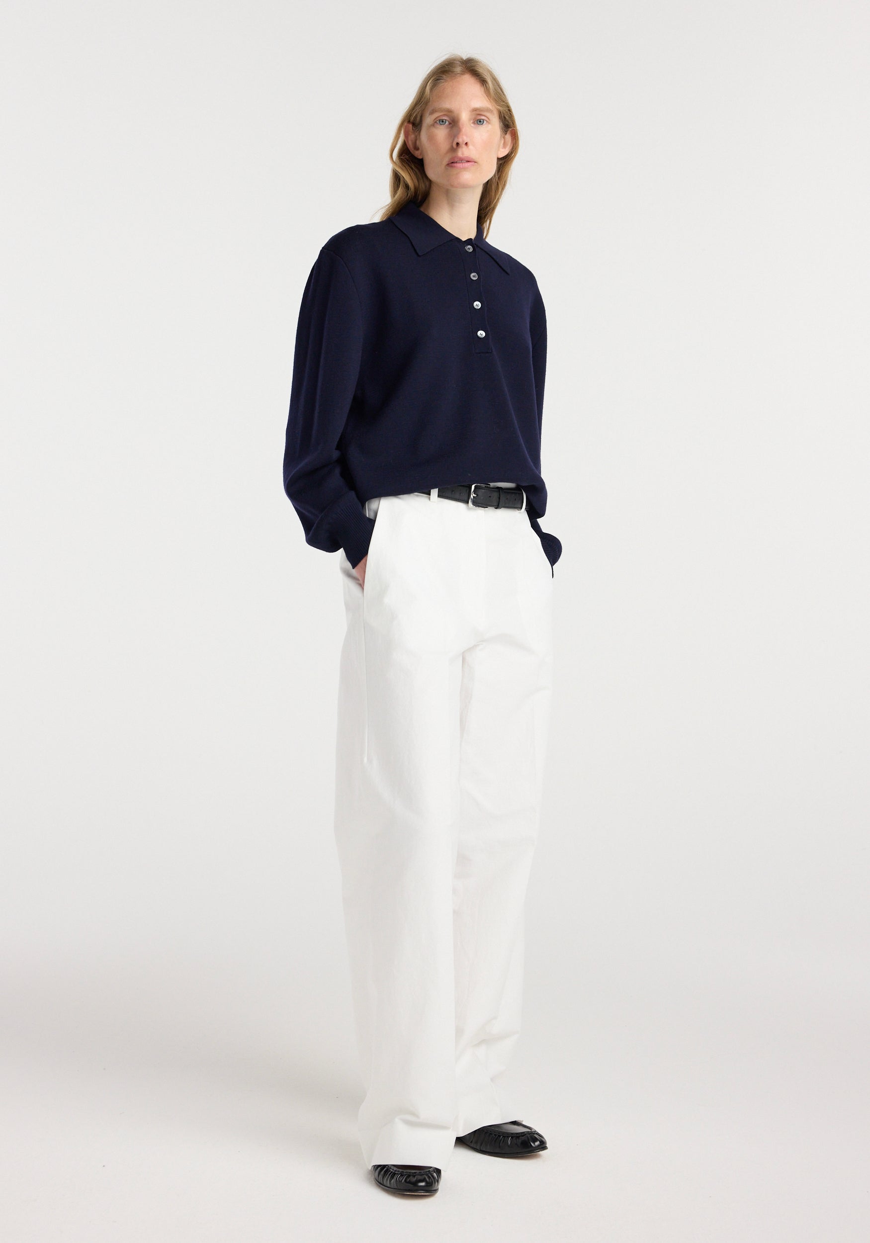 Woman wearing a navy Merino wool polo | navy with white high-waisted pants and black shoes, standing against a plain light background.