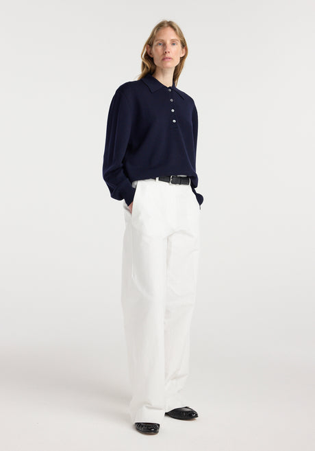 Woman wearing a navy Merino wool polo | navy with white high-waisted pants and black shoes, standing against a plain light background.