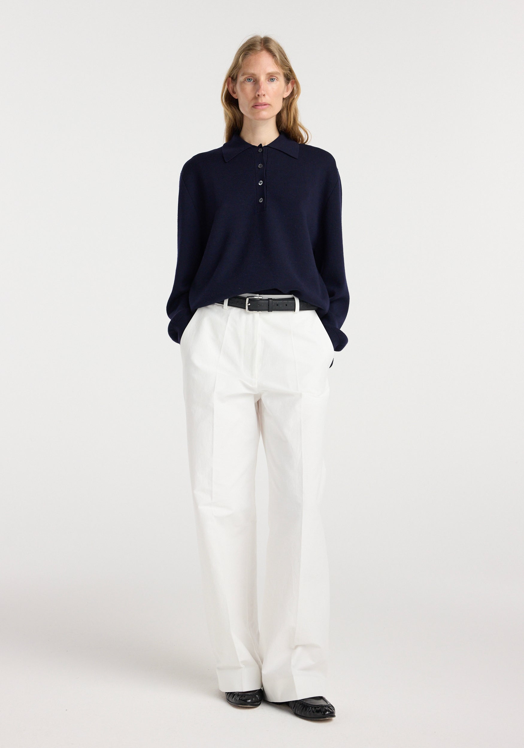 Woman wearing a navy Merino wool polo | navy with long sleeves, buttoned collar, tucked into white wide-leg pants with a black belt, standing against a plain background.