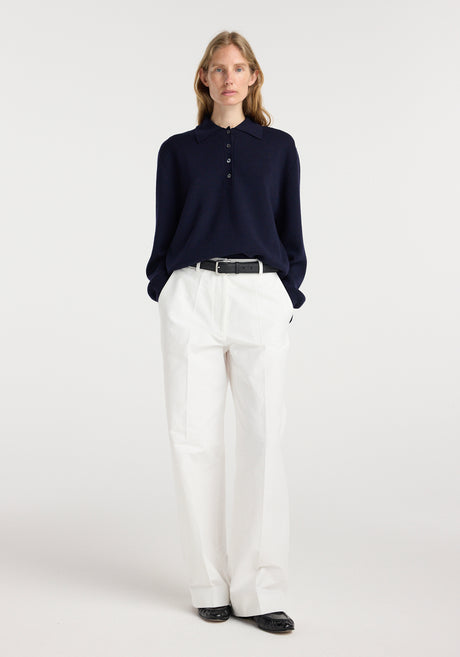 Woman wearing a navy Merino wool polo | navy with white high-waisted pants and black shoes, standing against a plain light background.