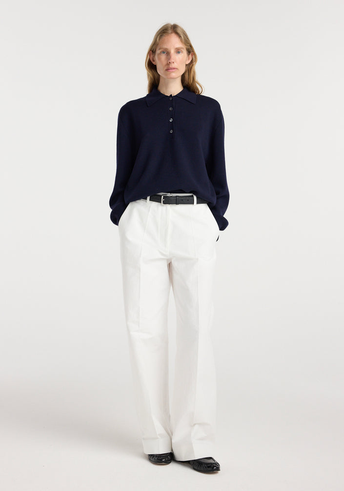 Woman wearing a navy Merino wool polo | navy with white high-waisted pants and black shoes, standing against a plain light background.