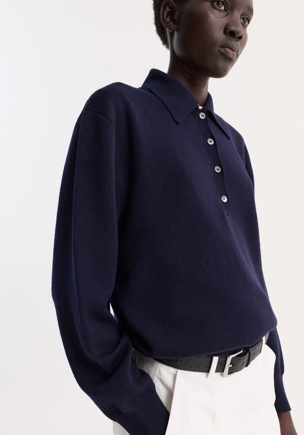Person wearing a Merino wool polo | navy, featuring a structured collar and three-button closure, tucked into high-waisted trousers, showcasing a relaxed, elegant style.