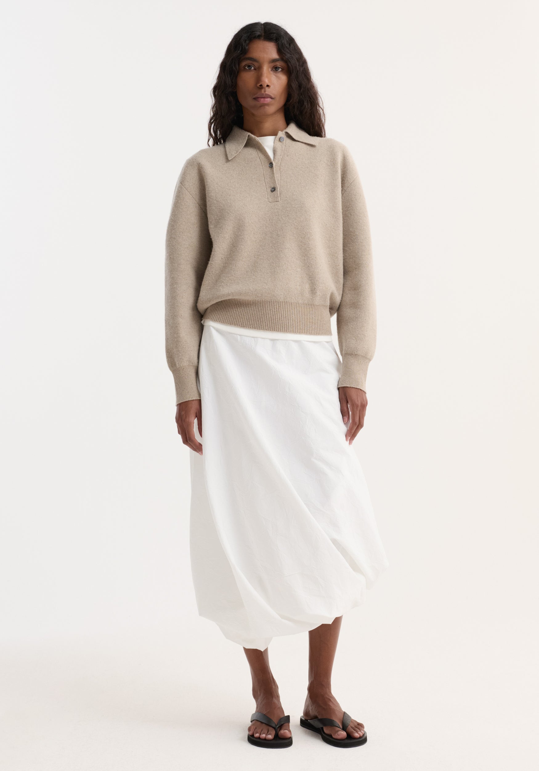 Person wearing Wool Cashmere Polo in stone melange, featuring a structured collar, three-button closure, paired with an ankle-length white skirt and black sandals.
