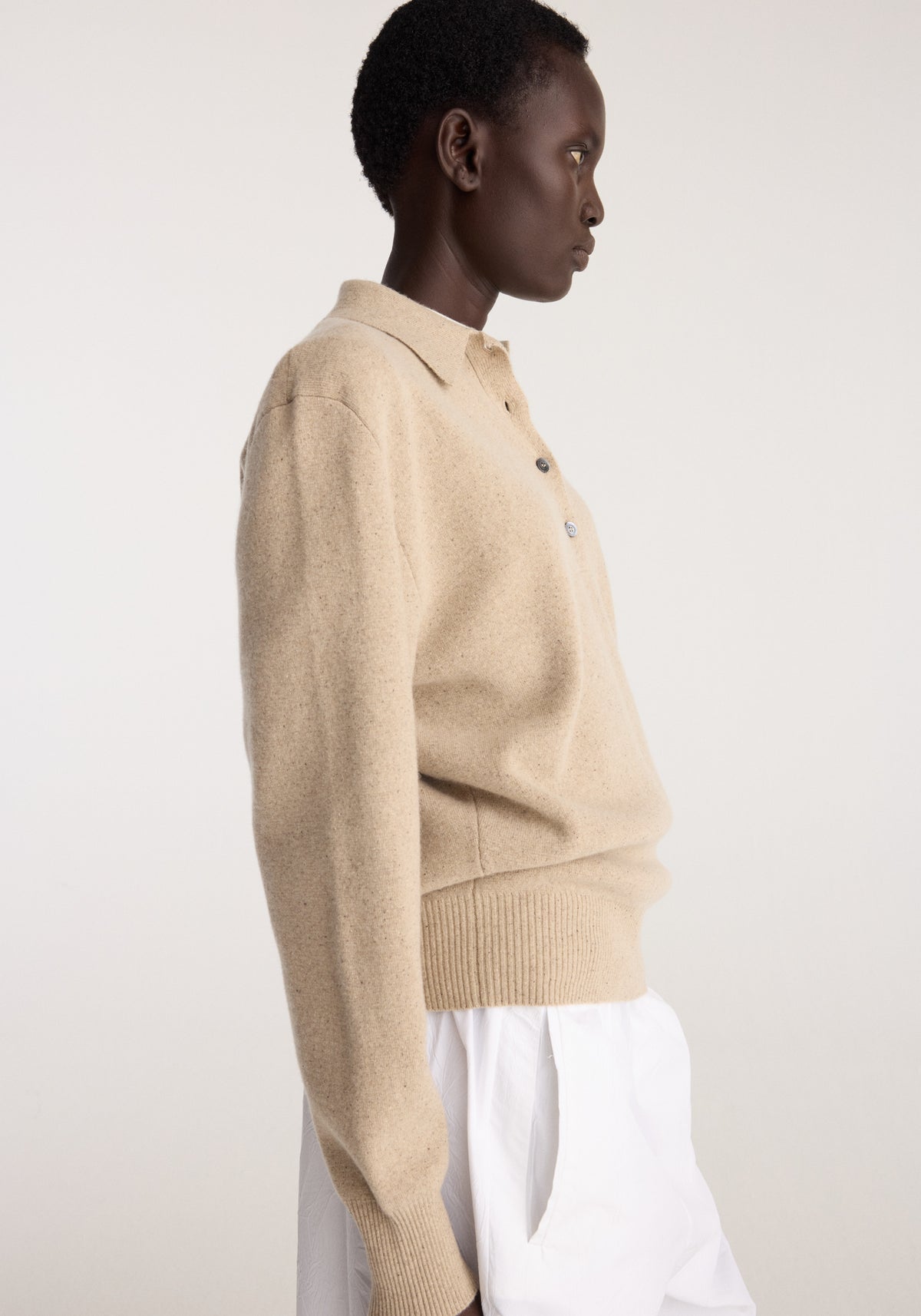 Side view of a model wearing Wool cashmere polo | stone melange in soft beige with ribbed cuffs and hem paired with white trousers