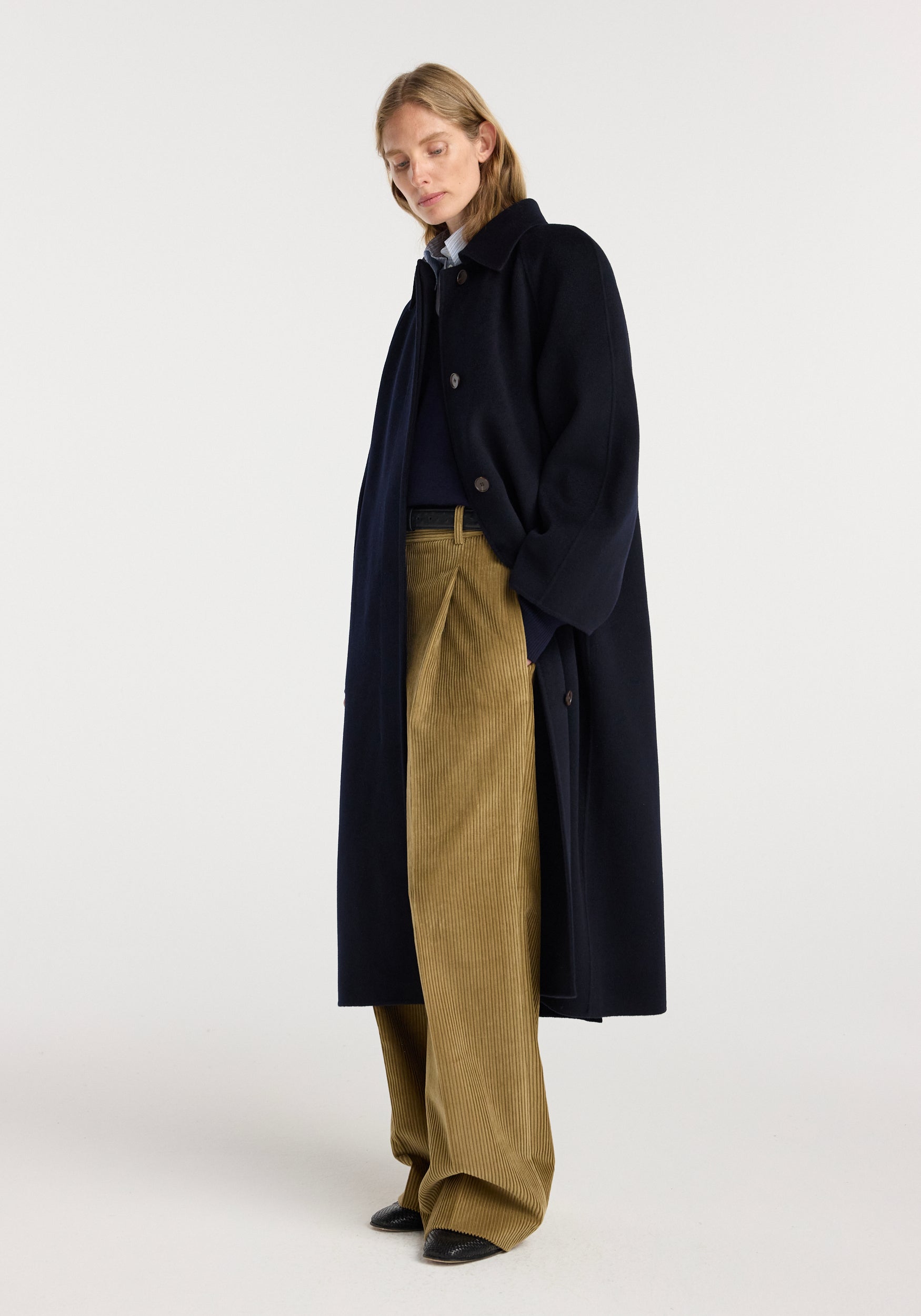 Woman wearing a long double-faced wool padded coat | navy with button closure, paired with beige wide-leg pants and black shoes, standing against a plain background.