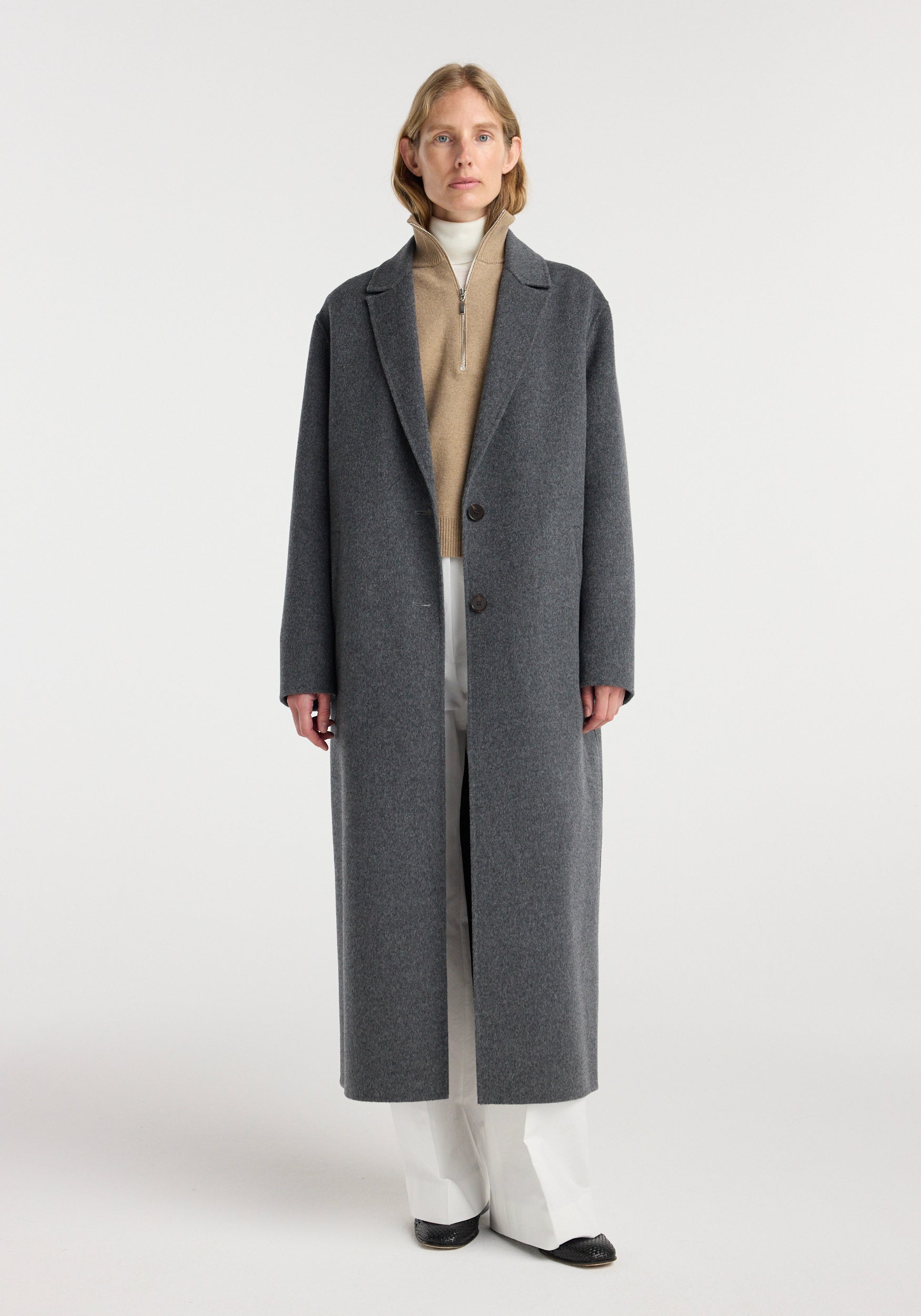Woman wearing a long dark grey melange double-faced wool long coat with a beige zip-up sweater, white pants, and black shoes standing against a plain background.