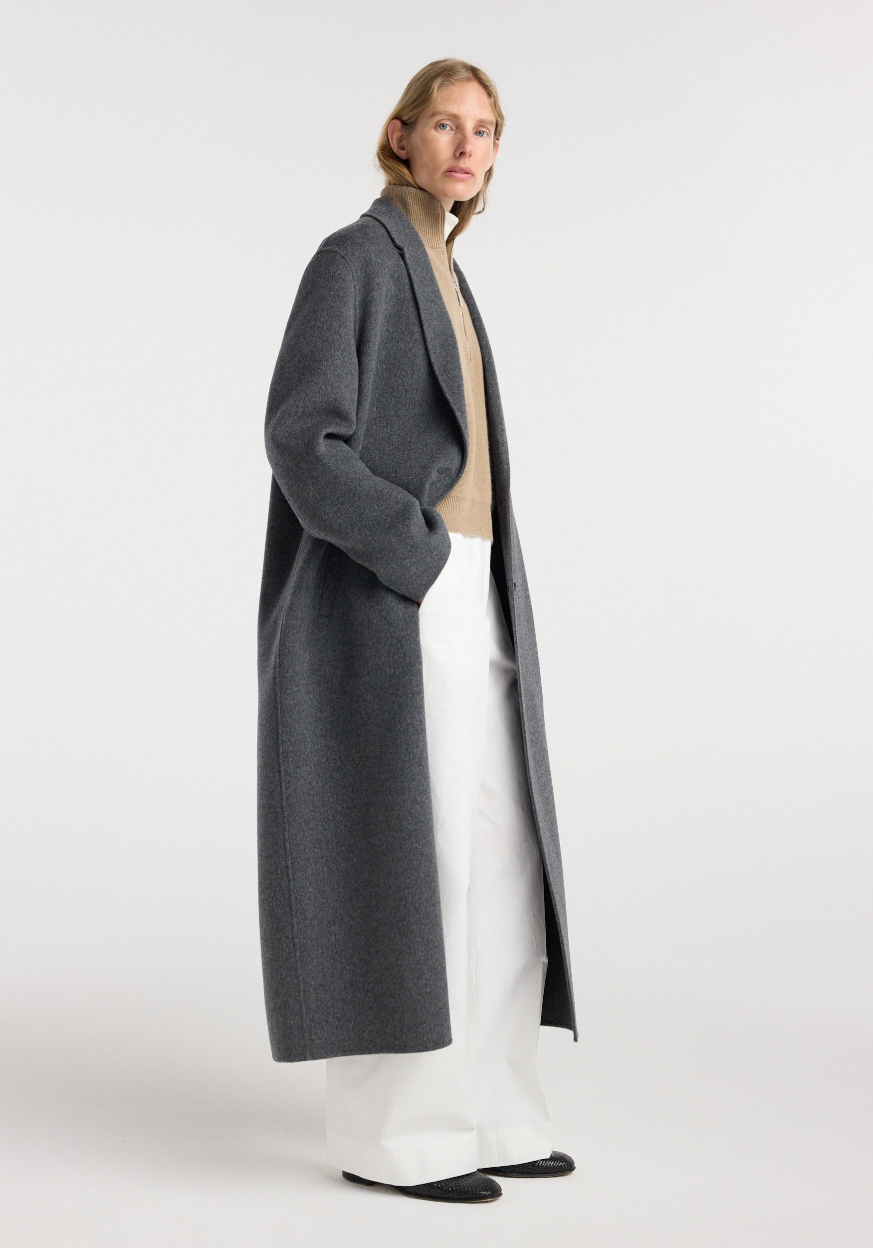Woman wearing a long dark grey melange wool coat, styled with a beige turtleneck sweater, wide white pants, and black shoes, standing against a plain background in the Double-faced wool long coat | dark grey melange.