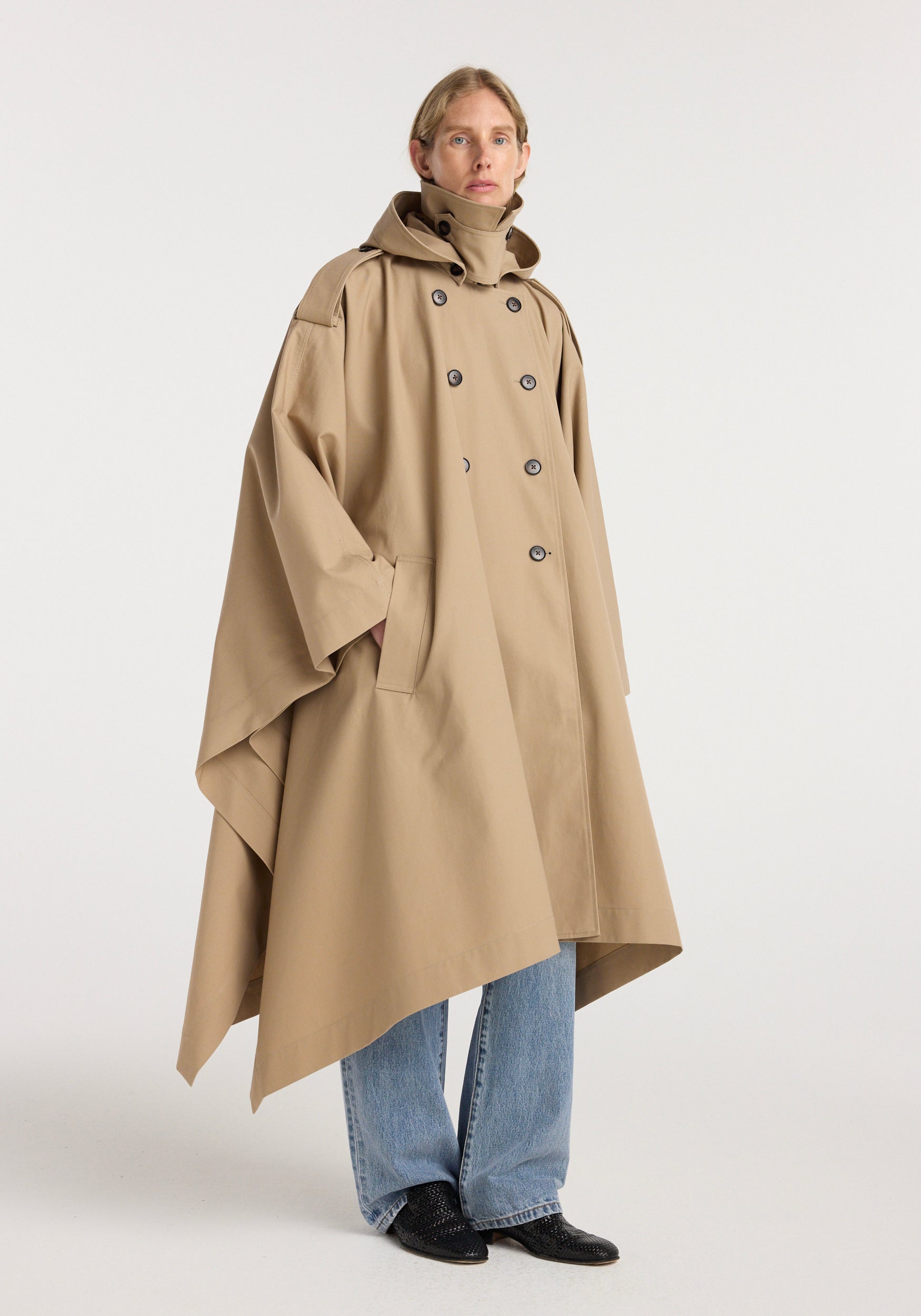 Model wearing a beige Hooded cape trenchcoat | beige with a double-breasted front, high collar, and side pockets, styled with blue jeans and black shoes.